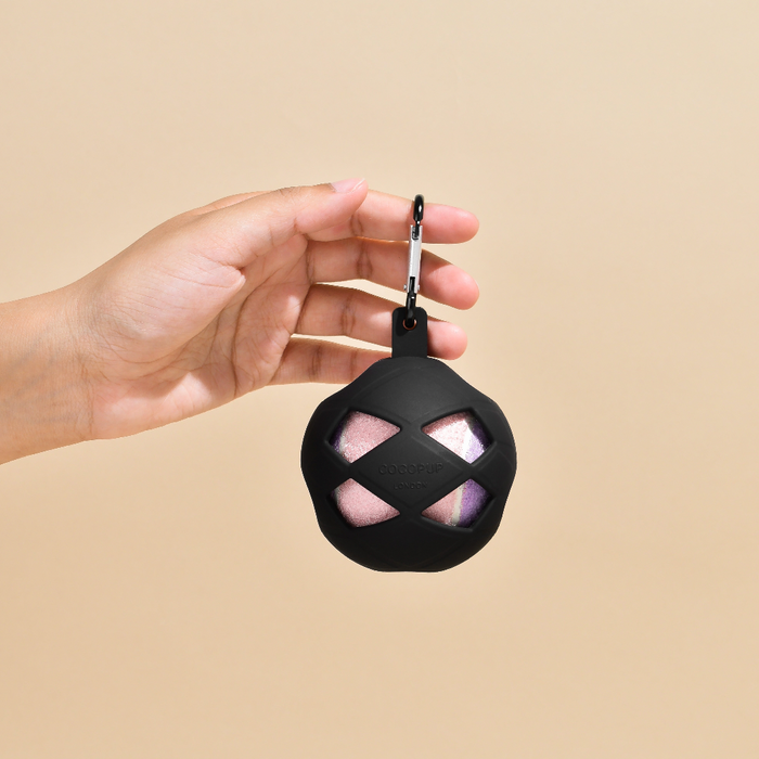 Hand holding Cocopup London Tennis Ball Holder with clip and pink tennis ball, shown against a beige background.