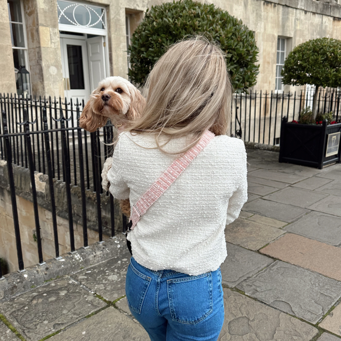 Woman with long blonde hair carries a dog and wears a pink textured tweed Cocopup London bag strap on a stone sidewalk.