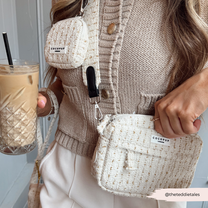Woman in a beige knit vest holds iced coffee and carries a cream textured tweed dog walking bag with matching dispenser.