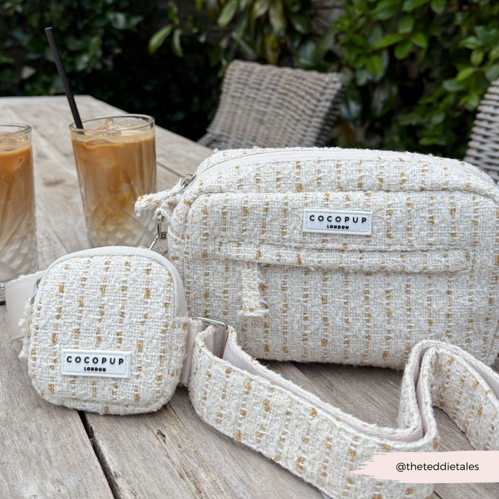 Vanilla textured tweed Cocopup London Dog Walking Bag on a wooden table with iced coffee, greenery behind.