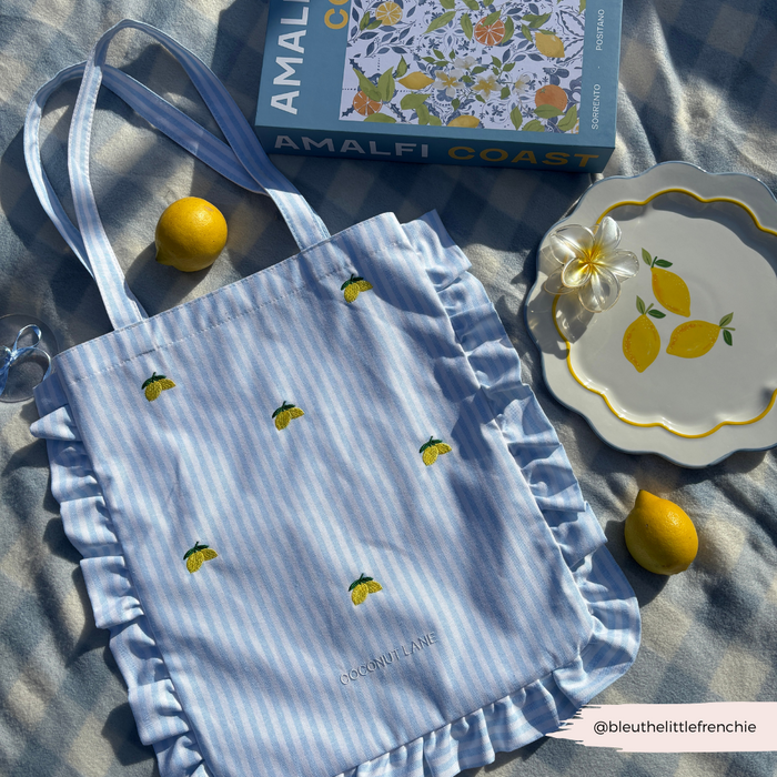 Yellow lemon-print tote bag and plate with lemons beside the Amalfi Coast book on a neutral blanket.