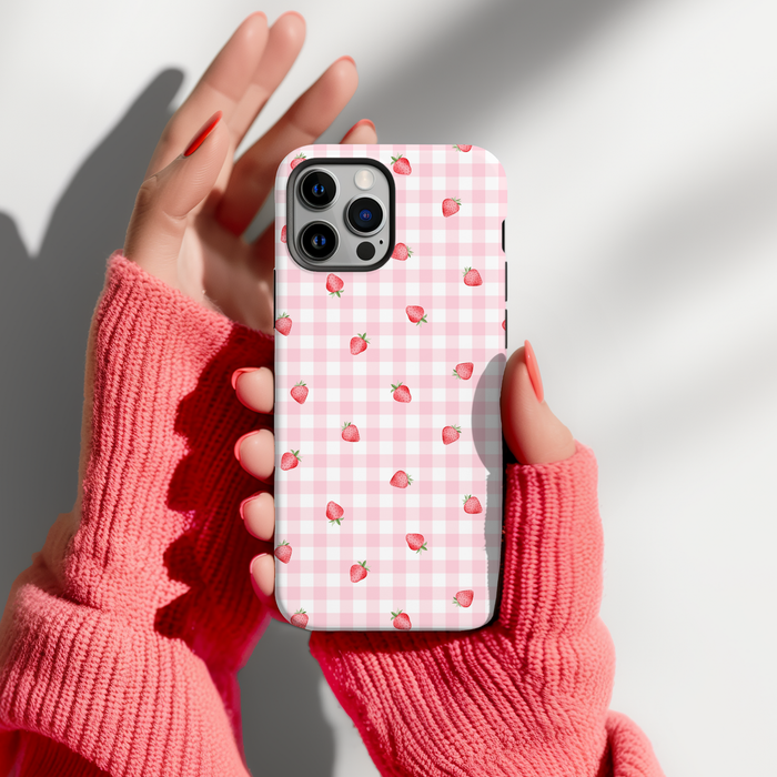 Hands in pink fingerless gloves hold a Cocopup London tough phone case with a pink strawberry and picnic blanket pattern.
