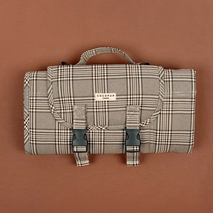 Folded Cocopup London plaid travel mat with buckle straps and handle, shown on a brown surface.