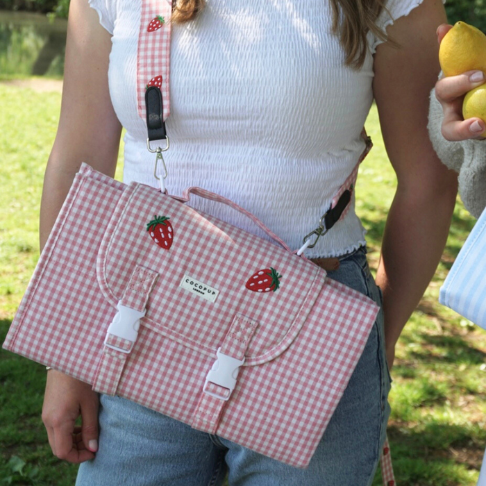 Pink gingham bag with strawberry patches held next to a Cocopup London travel mat in a matching strawberry pattern.