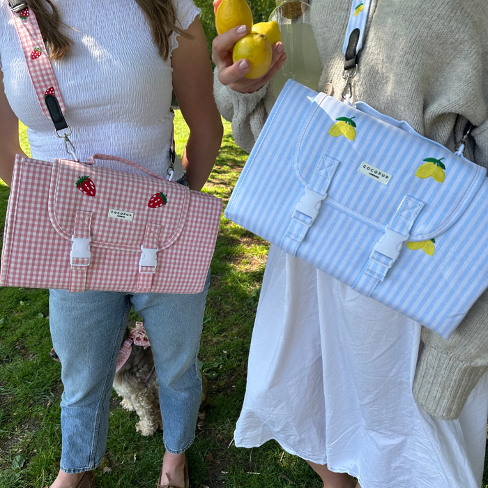 Two people outside with Cocopup London gingham handbags in pink strawberry and blue lemon prints, plus a picnic mat.