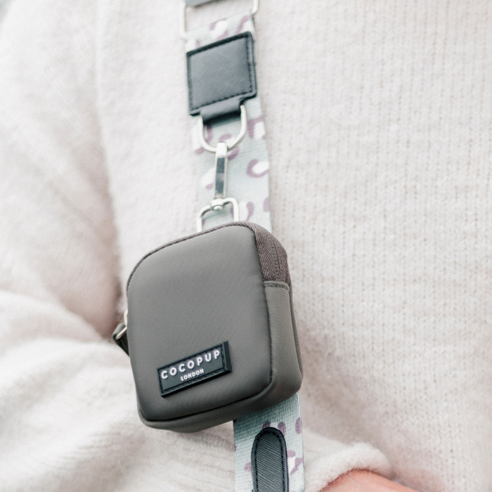 Close-up of a small, gray Treat Pouch - Mocha from Cocopup London attached to a light-colored, patterned strap. The strap features metal clips and leather details. The person wearing the mess-free bag is dressed in a soft, white sweater.