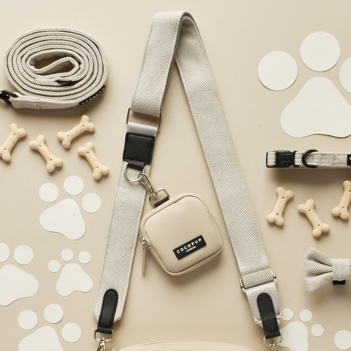 A flat lay image showcasing various beige and white dog accessories: an adjustable strap leash with a Cocopup London Tweed Bag Strap - Brown, a poop bag holder marked "DOG POOP," collar, bow tie, bone-shaped treats, and paw print cutouts. The items are arranged neatly on a neutral background.