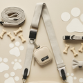 A flat lay image showcasing various beige and white dog accessories: an adjustable strap leash with a Cocopup London Tweed Bag Strap - Brown, a poop bag holder marked "DOG POOP," collar, bow tie, bone-shaped treats, and paw print cutouts. The items are arranged neatly on a neutral background.