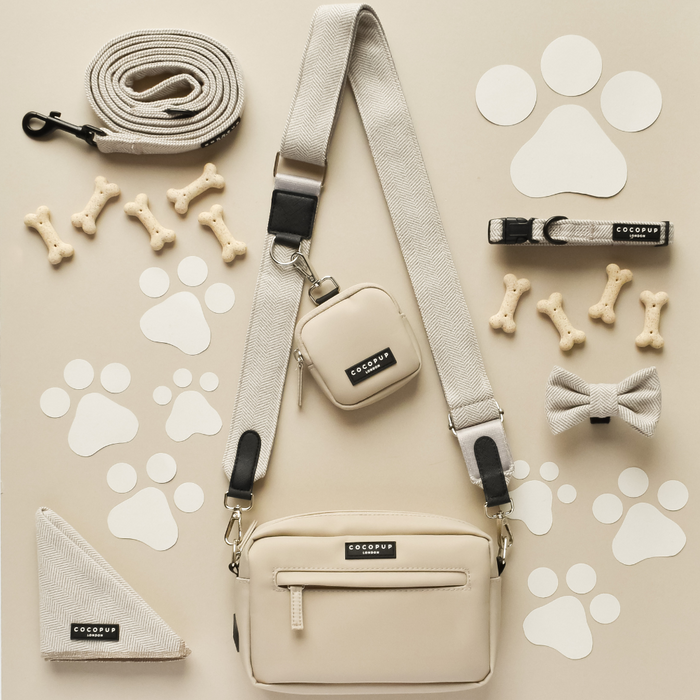 A flat lay of dog accessories, featuring a beige shoulder bag with a stylish Cocopup London Tweed Bag Strap in brown, leash, poop bag holder, collar, bow tie, bandana, and bone-shaped treats arranged on a light beige background with scattered white paw prints. The accessories are neatly organized and color-coordinated.
