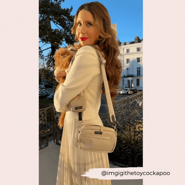 Woman in white with a small dog, wearing a brown Cocopup London tweed bag strap, outdoors near houses and trees.