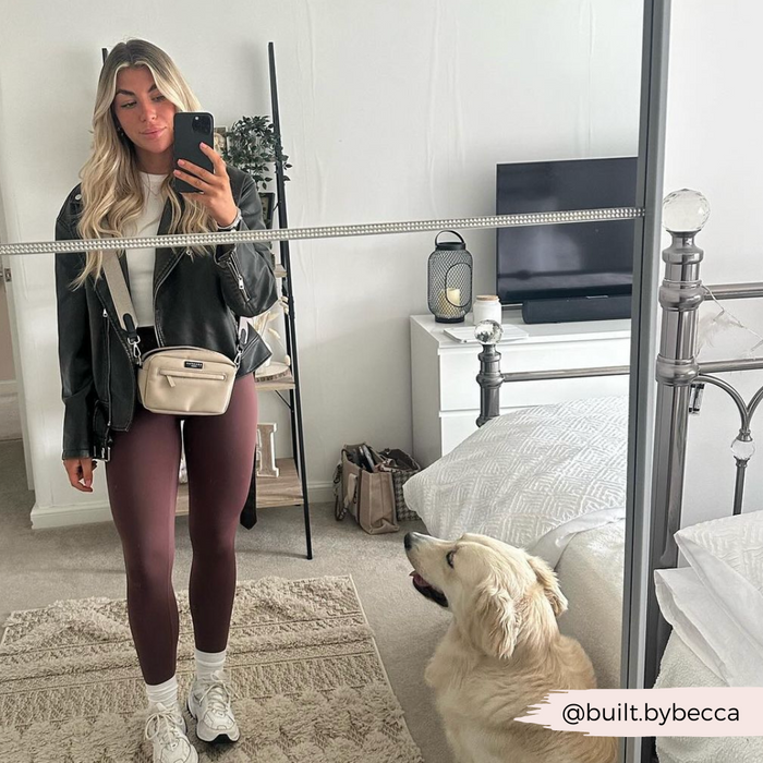 A woman with long blonde hair takes a mirror selfie in a bedroom. She is wearing a black jacket, white top, maroon leggings, and white sneakers. A golden retriever dog is sitting on the floor, looking up at her. The Instagram handle "@built.bybecca" is in the bottom right corner and an accessory from Cocopup London featuring the Tweed Bag Strap - Brown rests nearby.