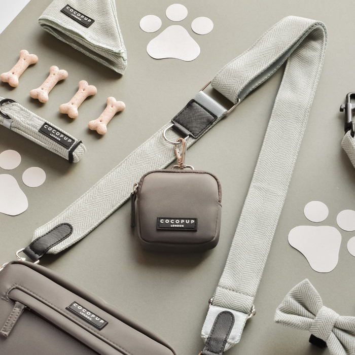 A flat lay photo features pet accessories, including a grey leash, waste bag holder, collar, and personal pouch from "Cocopup London." Also in the image are dog bone-shaped treats, a small bow tie, and paw print designs on a light green background with the stylish Tweed Bag Strap in Sage from Cocopup London – perfect for your dog's walking bag.