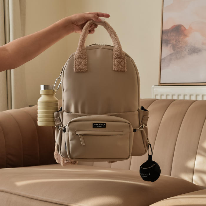 Caramel Latte rucksack with teddy handles, bottle clip, and ball holder, displayed on a tan sofa.