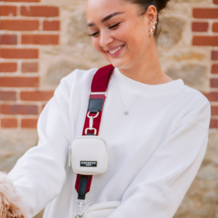 Woman in a white sweatshirt with a berry red velvet bag strap, petting a dog beside a brick wall.