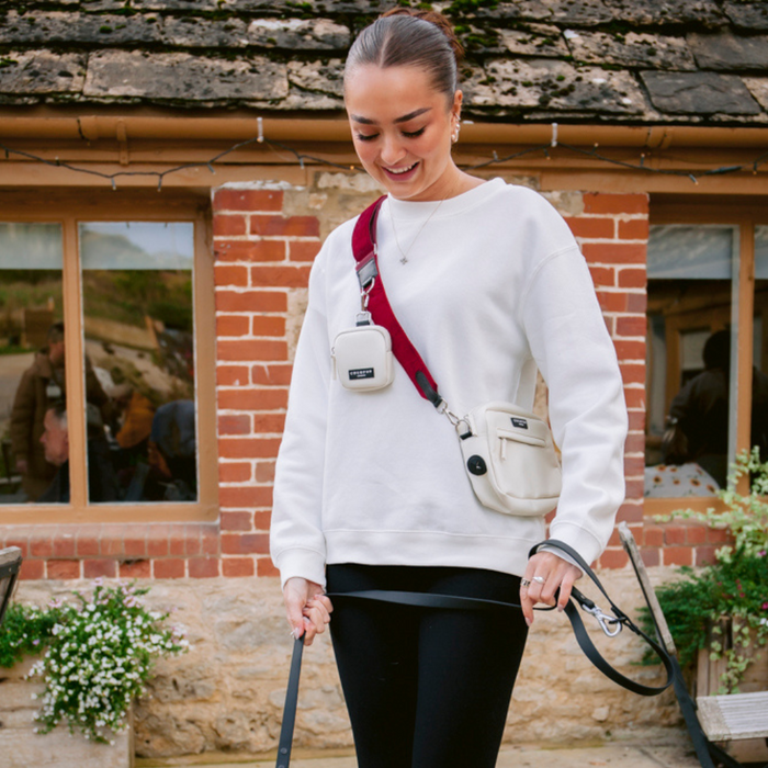 Woman in white sweatshirt holds dog leash with Cocopup London berry red velvet bag strap outdoors.