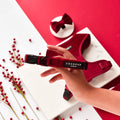 Hand holding a berry red velvet dog collar on a white surface, with scattered berries and a bow tie nearby.