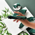 Hand holding a forest green velvet dog collar with gold hardware; matching bowtie, leash, and green leaves nearby.