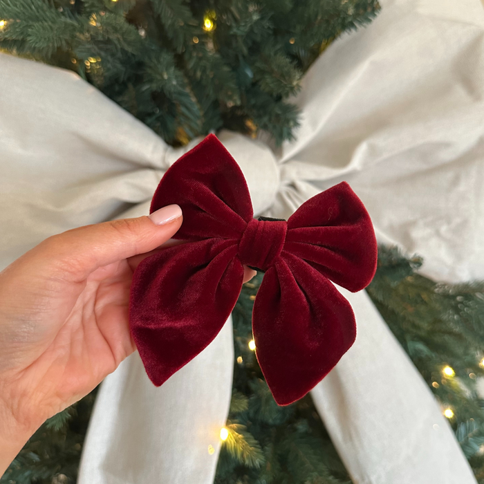 Hand holding a velvet berry red Cocopup London sailor bow tie with a ribbon-wrapped, lit Christmas tree in background.