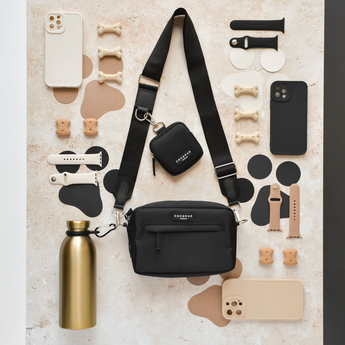 Black bag, Cocopup London Water Bottle Holder Clip, smartwatches, phone cases, and dog bone treats on marble surface.