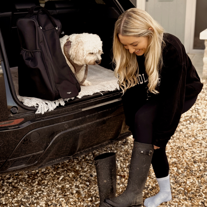 Woman putting on tall boots in a car with her dog next to a black Cocopup London welly boot bag.