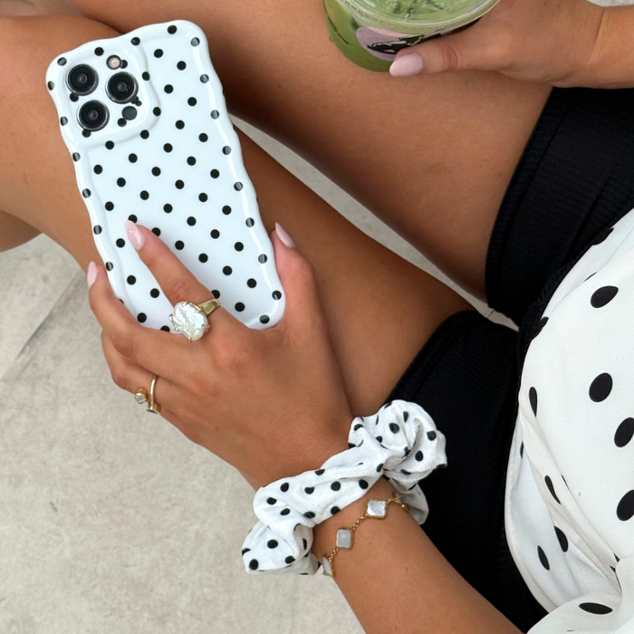 Woman with a white scrunchie with black polka dots in her hair, holding a green drink and phone.